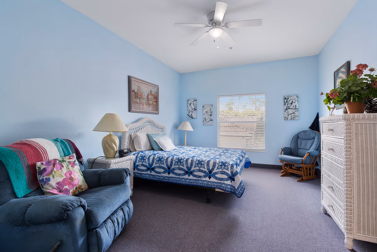A cozy bedroom with light blue walls and a ceiling fan. The room features a bed with a blue and white patterned quilt, a nightstand with a lamp, a blue upholstered armchair with a colorful blanket and floral pillow, a rocking chair, and a white wicker dresser with a potted plant on top. There is a window with blinds letting in natural light and artwork hanging on the walls.