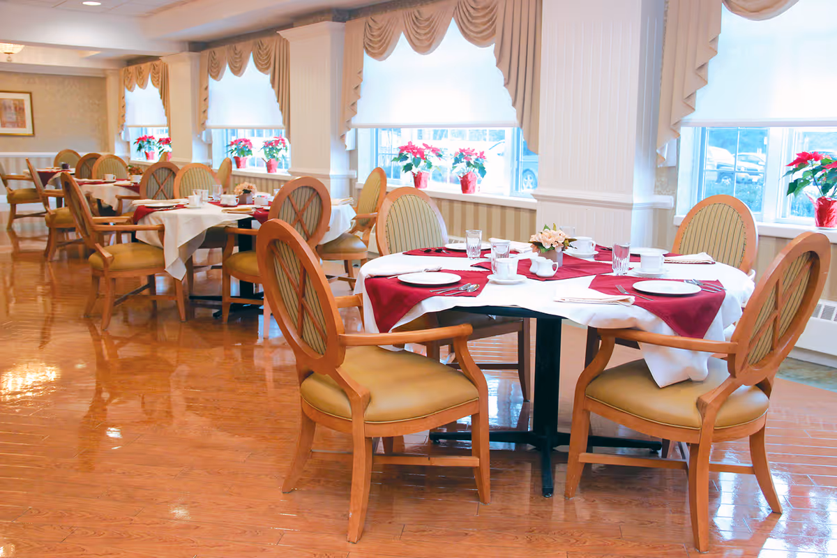 Bright dining room with round tables set for a meal, wooden chairs, and poinsettias on the windowsills.