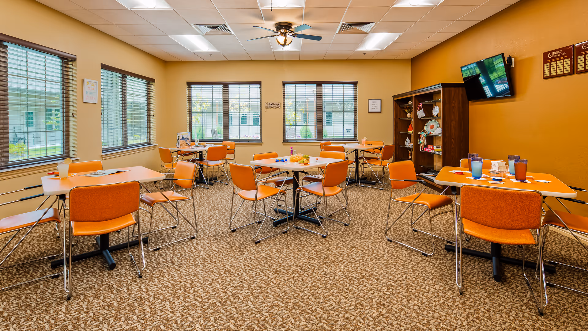 Bright communal dining/activity room with orange chairs and tables, carpeted floor, windows along the back wall, and a TV mounted on the side.