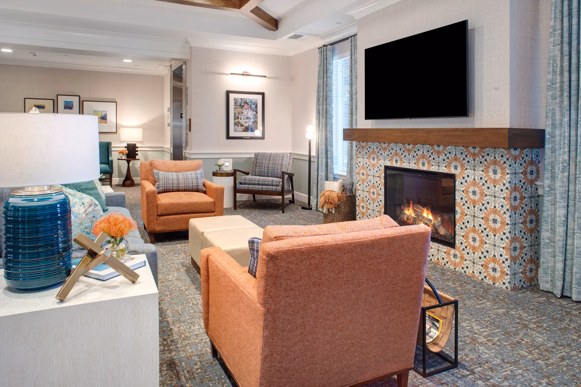 A cozy living room with orange armchairs, a patterned fireplace topped by a wall-mounted TV, a lamp on a side table, and additional seating in the background.