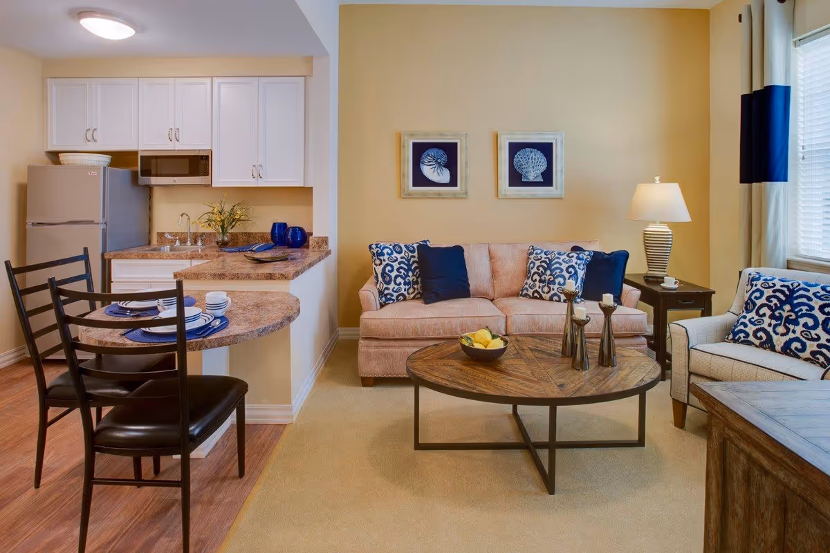 A cozy living area with a beige sofa adorned with blue and white patterned pillows, a matching armchair, and a round wooden coffee table with decorative items. To the left is a small kitchen area with white cabinets, a refrigerator, microwave, and a countertop with two place settings and chairs. The walls are painted a warm yellow, and there are two framed shell artworks above the sofa. A lamp sits on a side table next to the sofa, and a window with curtains allows natural light into the room.