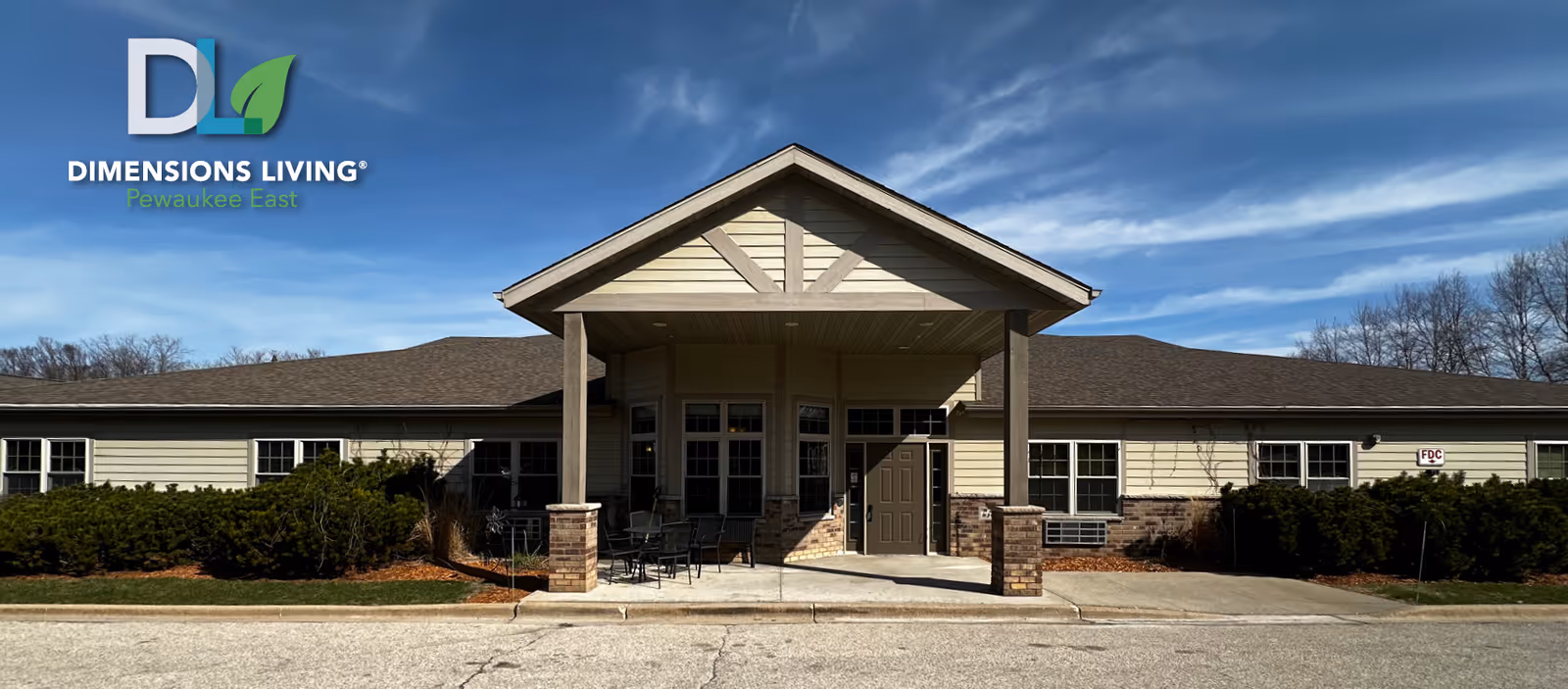 Exterior front view of Dimensions Living Pewaukee East facility showing a single-story building with a covered entrance supported by four columns, outdoor seating area, and landscaping with bushes under a partly cloudy blue sky.