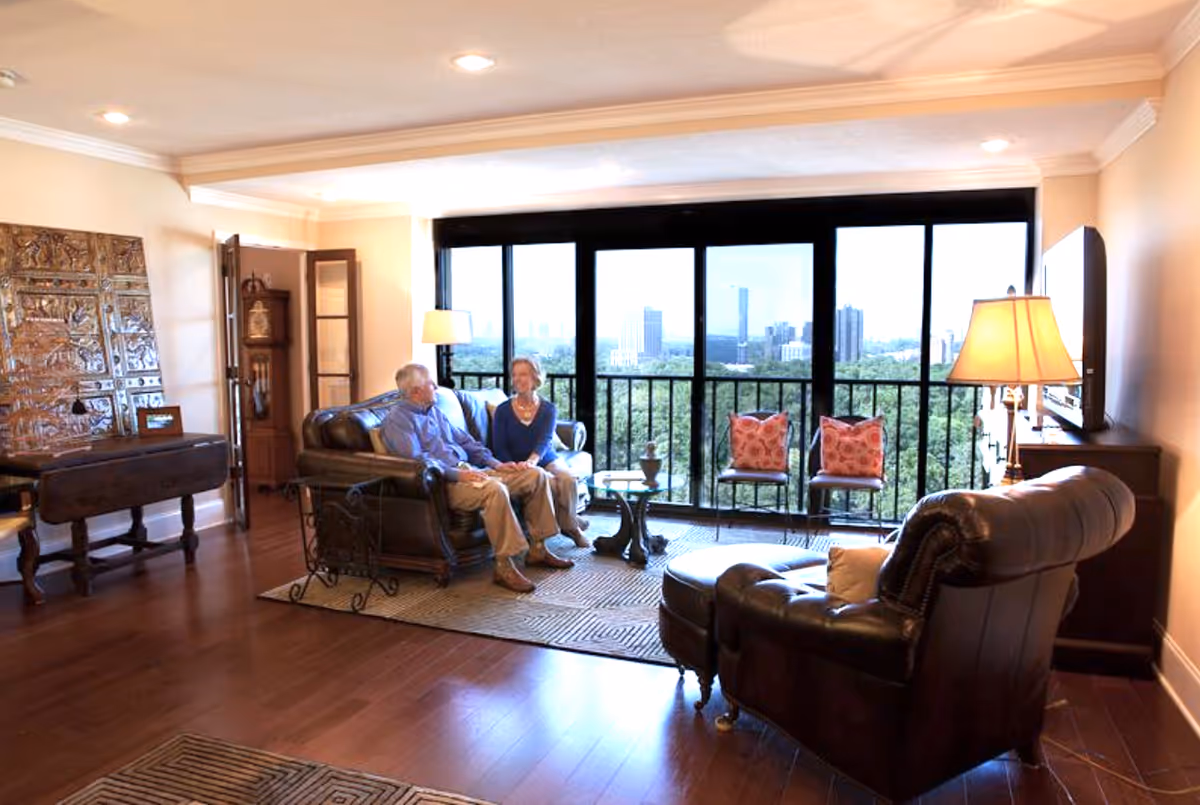 Two people sit on leather sofas in a bright living room with large windows overlooking a city skyline.