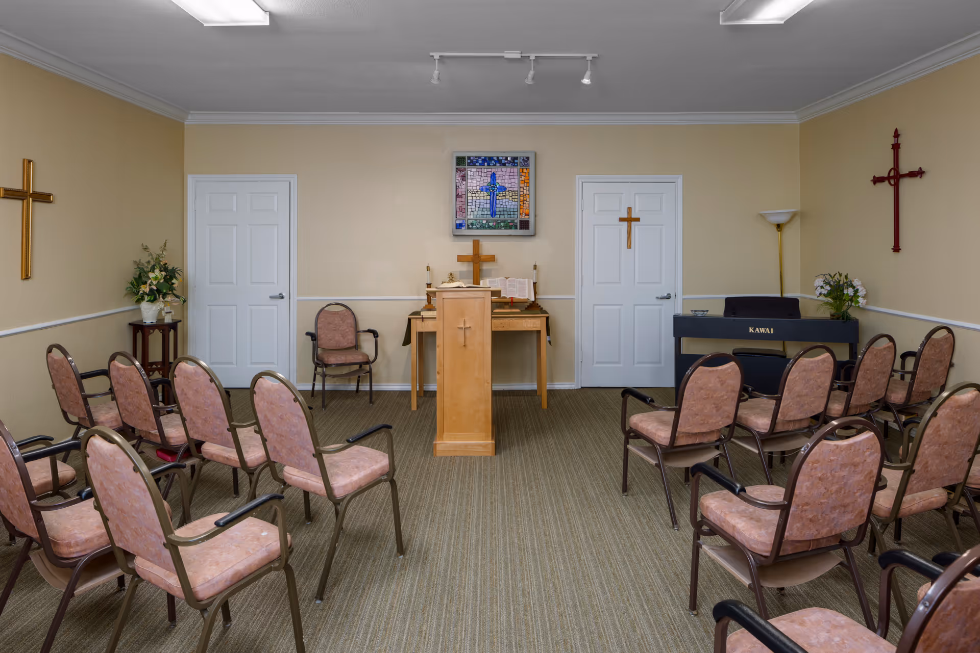 A small chapel or prayer room with rows of pink cushioned chairs facing a wooden podium with a cross. Behind the podium is a table with an open Bible and a cross, flanked by two closed white doors. The walls are beige with three crosses mounted on them and a stained glass artwork featuring a cross above the table. There is a black Kawai keyboard on the right side and floral arrangements on stands.