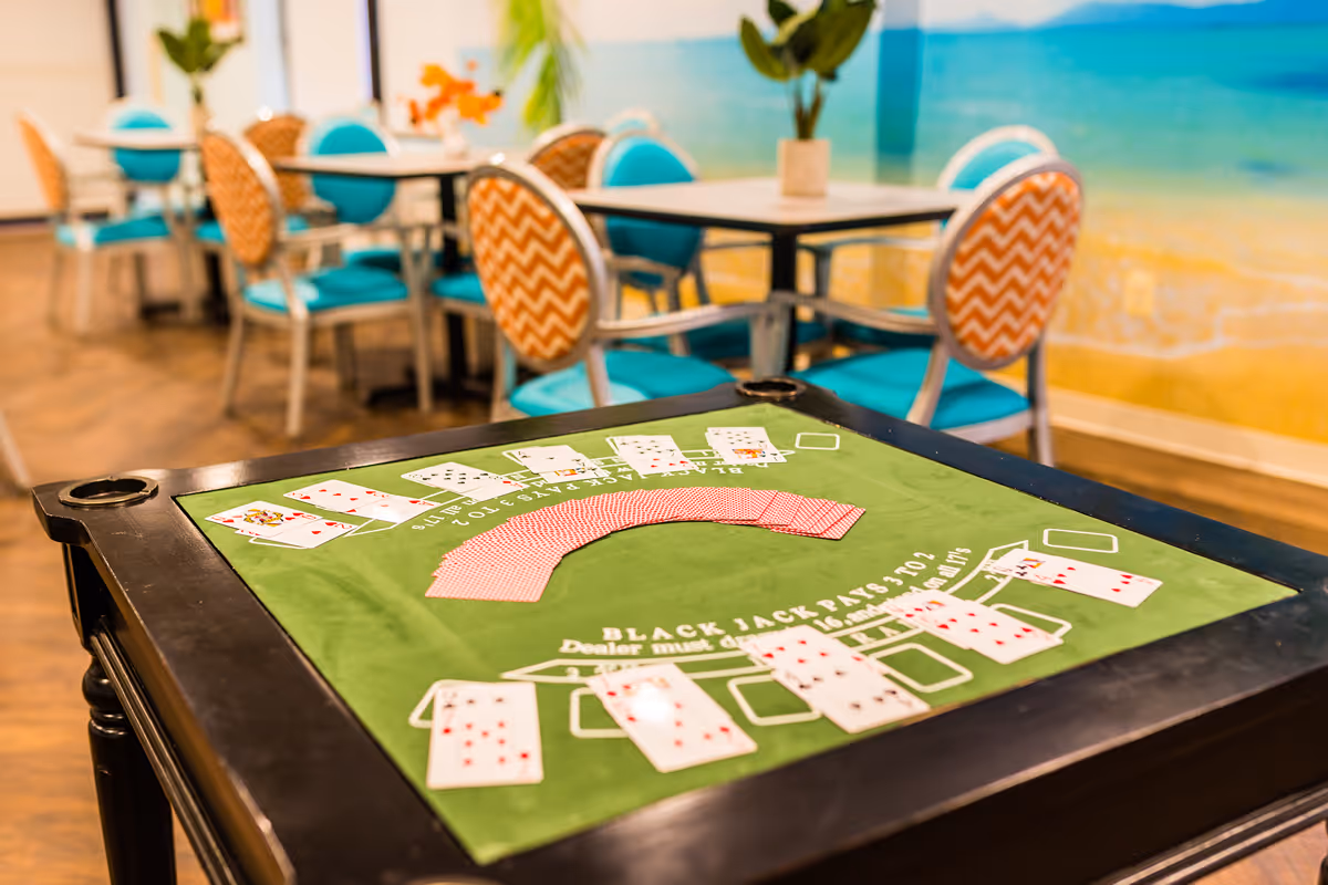 A blackjack card table with playing cards laid out, situated in a room with tables and chairs. The chairs have turquoise cushions and orange chevron-patterned backs. The background features a wall mural depicting a beach scene with sand and ocean.