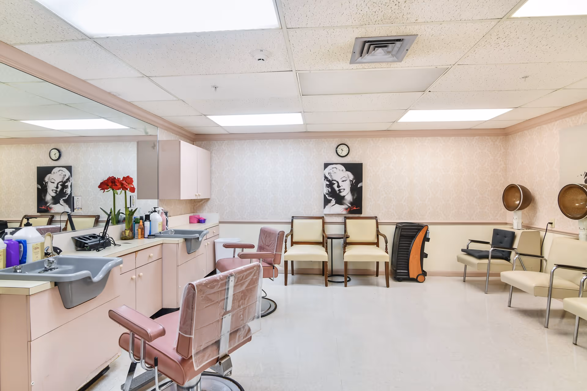 Interior of a hair salon room with pink salon chairs, sinks, and various hair care products on the counter. There are two vintage hair dryers on the right side, cream-colored chairs along the wall, a small table between two chairs, and a black and white portrait of Marilyn Monroe on the wall under a clock.