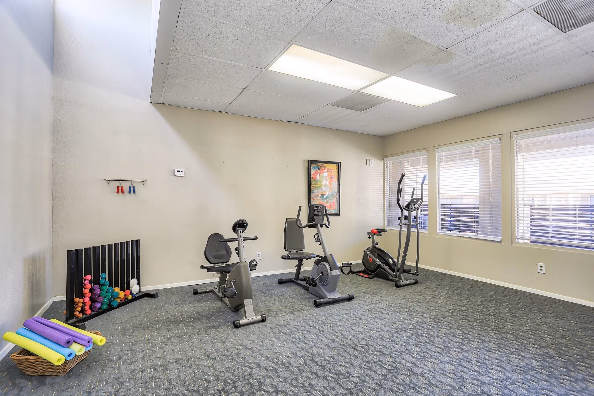 A small fitness room with exercise equipment including two recumbent stationary bikes, an elliptical machine, a rack of colorful dumbbells, and a basket of foam rollers. The room has beige walls, a carpeted floor, fluorescent ceiling lights, and three windows with blinds letting in natural light.