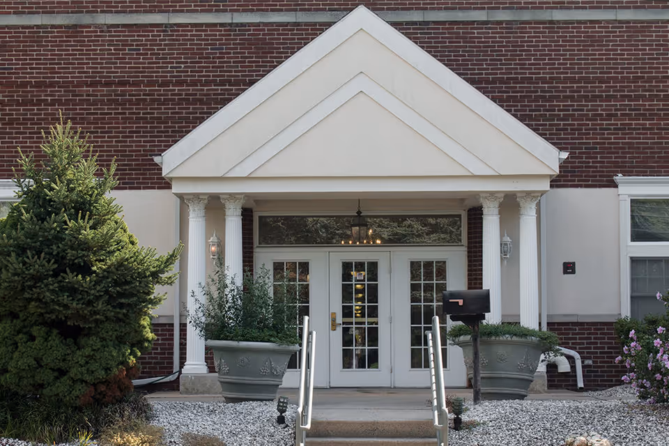 Entrance of a brick building with white double doors and a white triangular pediment supported by four white columns. There are two large planters with greenery on either side of the entrance, a black mailbox on the right, and a small set of stairs with handrails leading up to the doors.