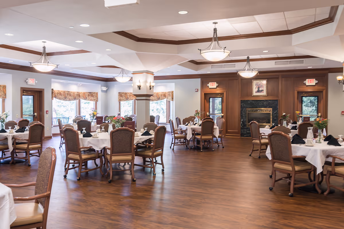 A spacious dining room with multiple round tables covered with white tablecloths and set with glassware and napkins. The room features wooden floors, cushioned chairs, large windows with patterned valances, and a fireplace with a framed picture above it. Ceiling lights and wall sconces provide warm lighting.