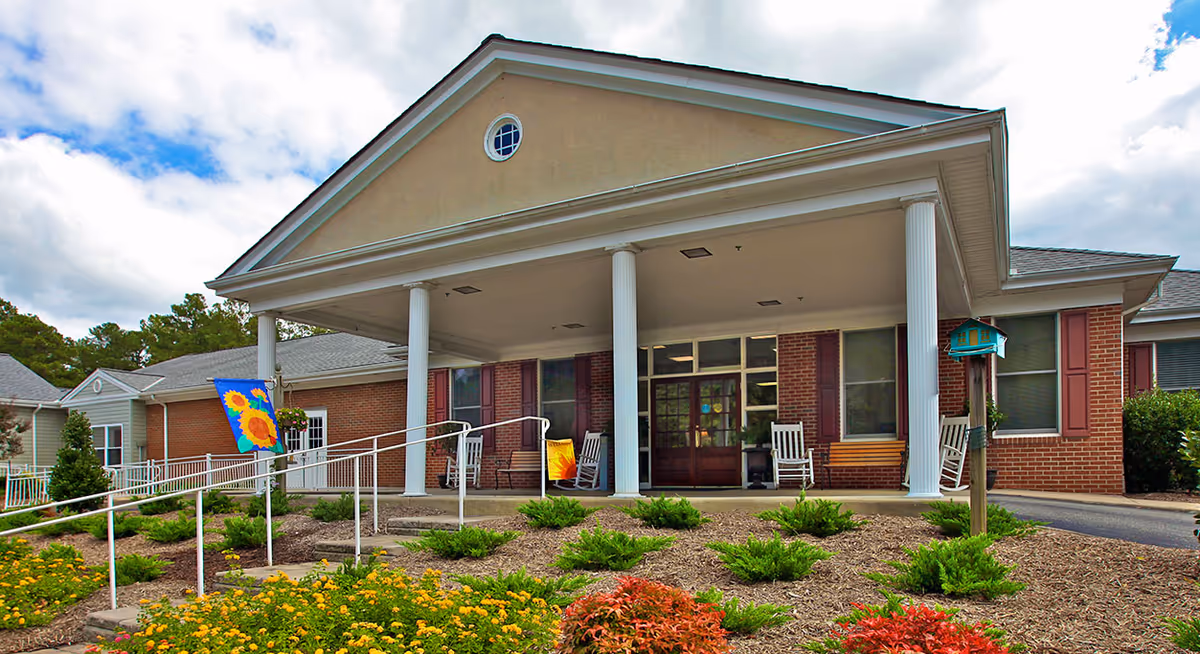 Covered front entrance of a senior living building with white columns, a wheelchair ramp, rocking chairs, and landscaped flowerbeds.