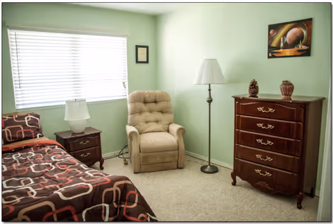 A small green-walled bedroom with bed, dresser, chair, and lamps.