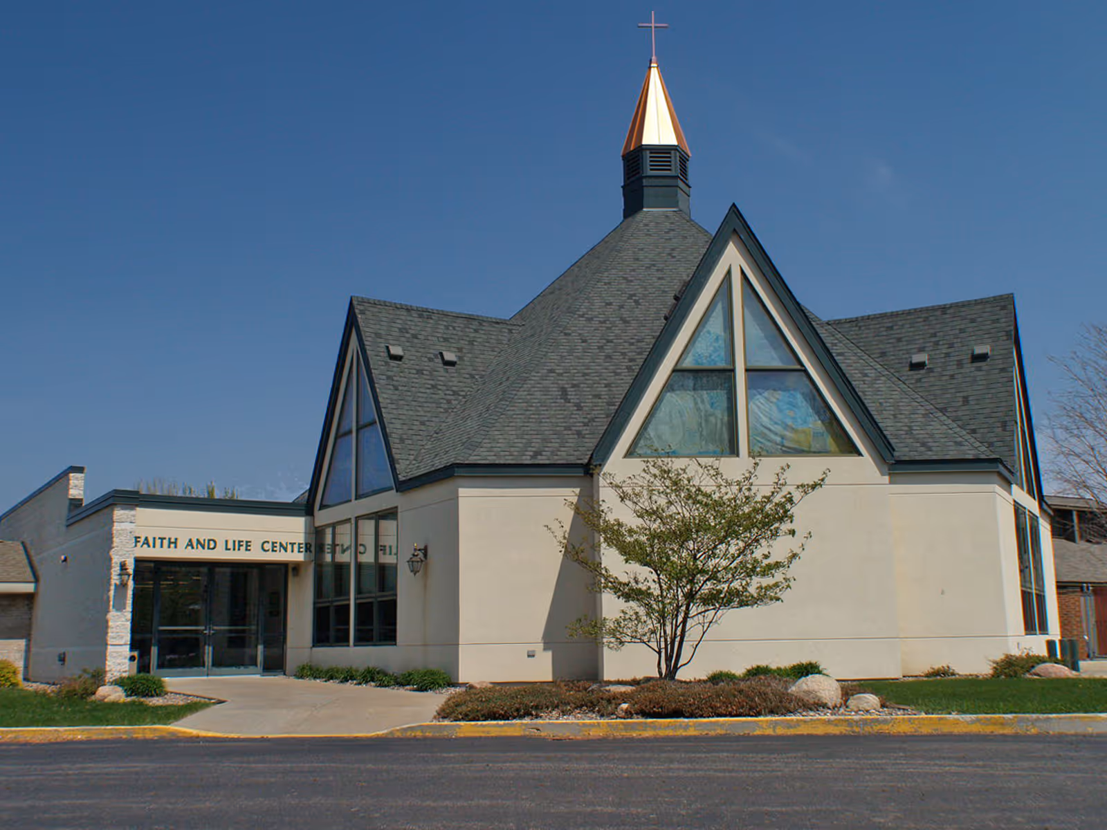 Exterior view of a building with a steeply pitched roof and a small steeple topped with a cross. The building has large triangular windows with stained glass and a sign that reads 'Faith and Life Center' above the entrance. There is a small tree and landscaping in front of the building under a clear blue sky.
