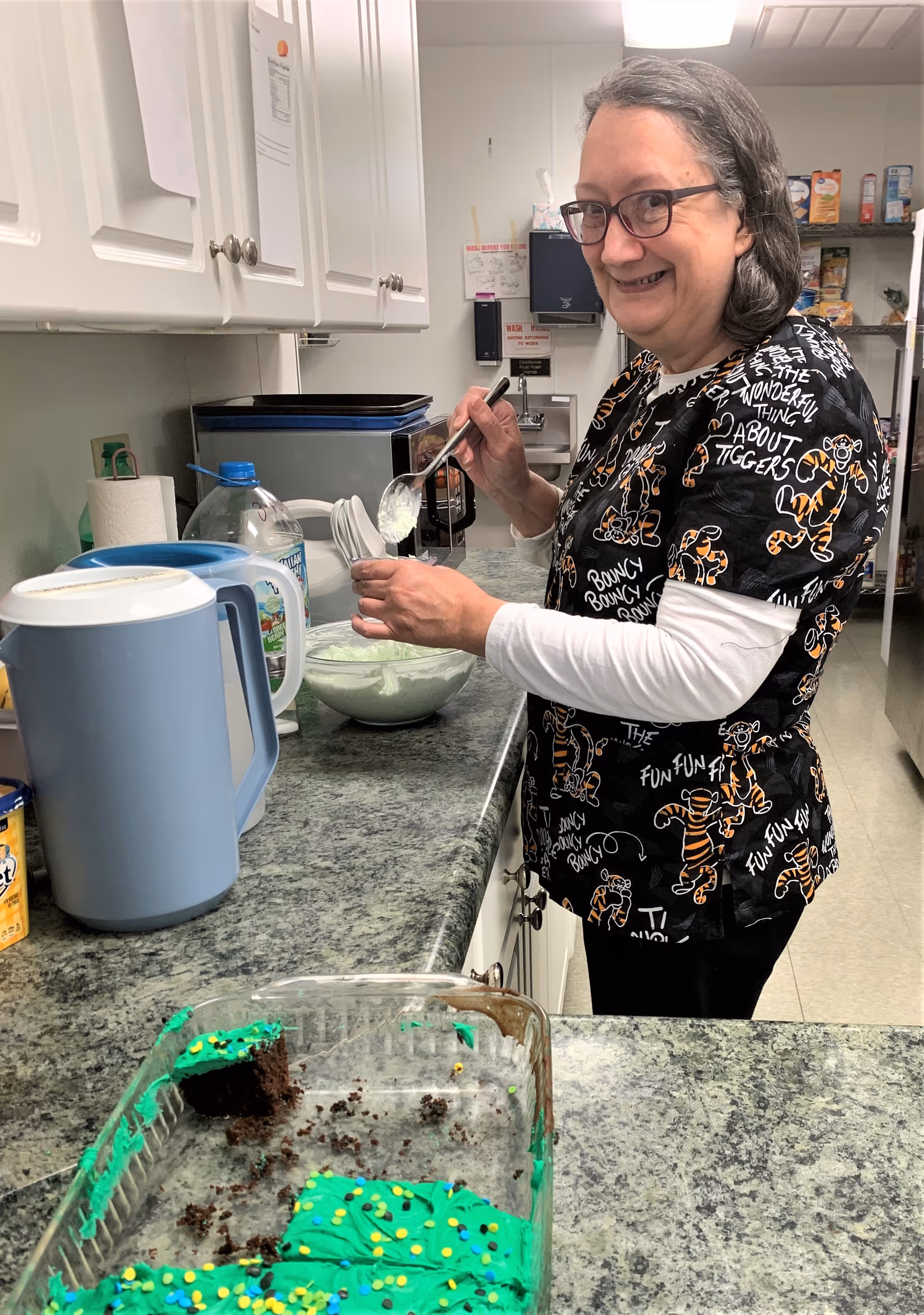 A woman wearing glasses and a black shirt with orange tiger prints is smiling while scooping white frosting from a bowl in a kitchen. In the foreground, there is a partially eaten chocolate cake with green frosting and colorful sprinkles on a countertop. The kitchen has white cabinets, a blue pitcher, and various items on shelves in the background.