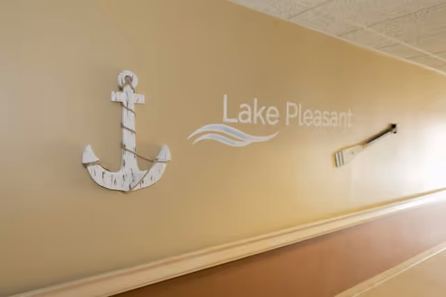 A beige hallway wall decorated with a white wooden anchor on the left, the words 'Lake Pleasant' in white with a wave design in the middle, and a wooden oar mounted on the right side.