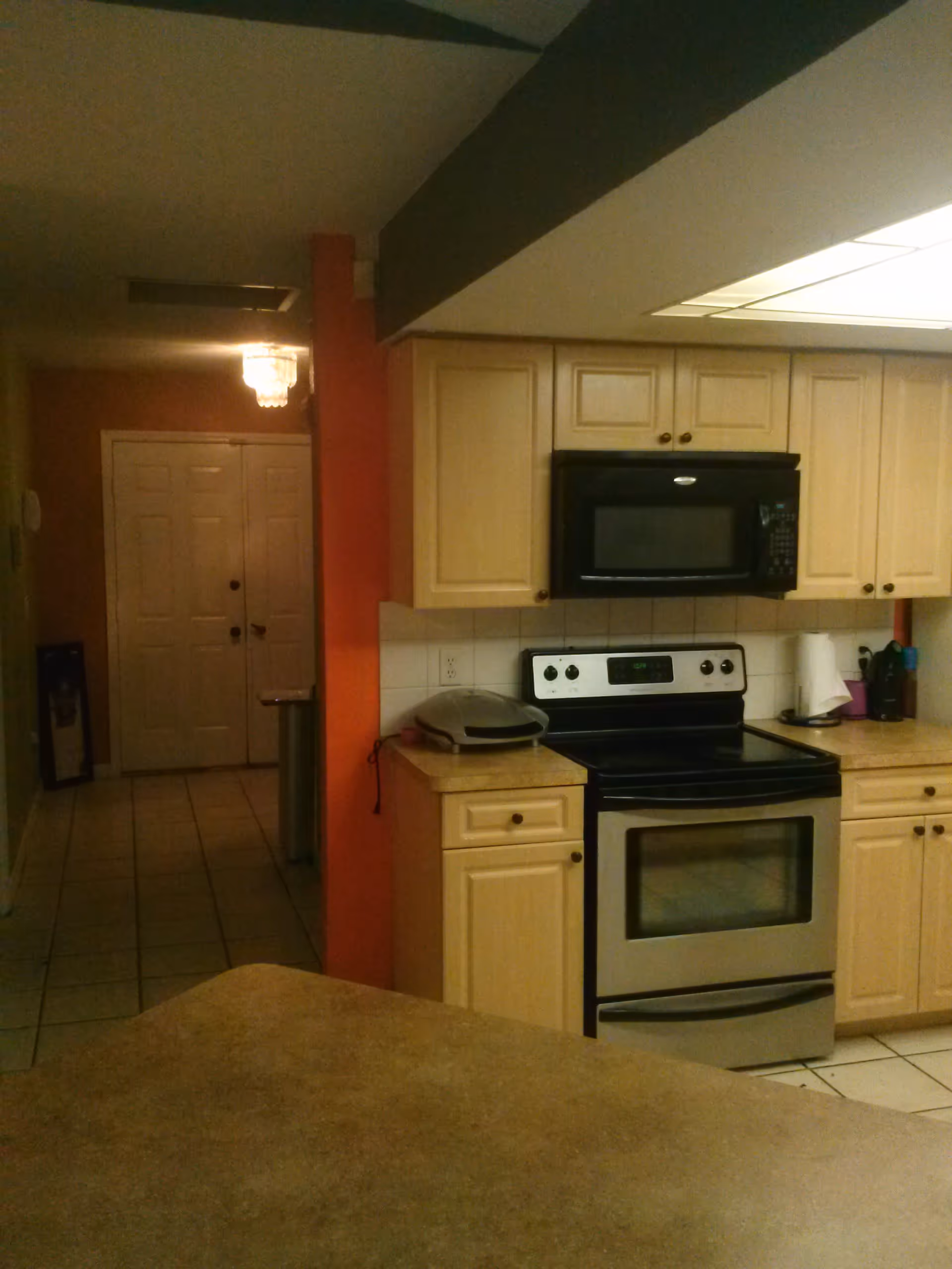 Interior view of a kitchen with light wood cabinets, a black microwave above a stainless steel electric stove, and a countertop with a small appliance and paper towel holder. The kitchen opens to a hallway with double doors and a chandelier light fixture.