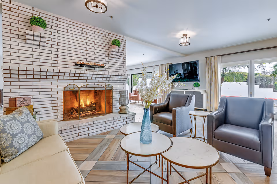 A cozy living room with a lit brick fireplace, beige sofa with patterned cushions, two dark leather armchairs, and three round marble-top coffee tables with a blue vase holding white flowers. The room has large windows with beige curtains, a wall-mounted TV, and decorative green plants on the fireplace and shelves.