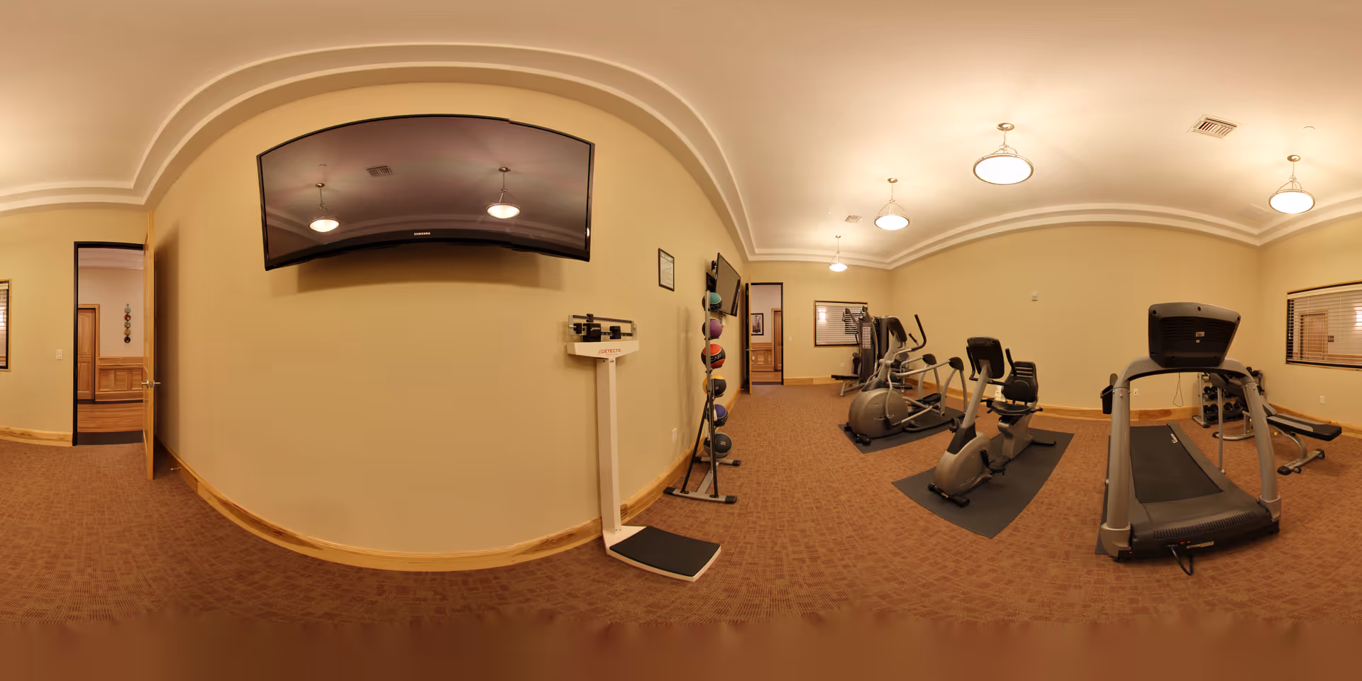 Panoramic gym/fitness room with treadmills, stationary bikes, free weights, a wall-mounted TV, and a scale in an assisted living facility.