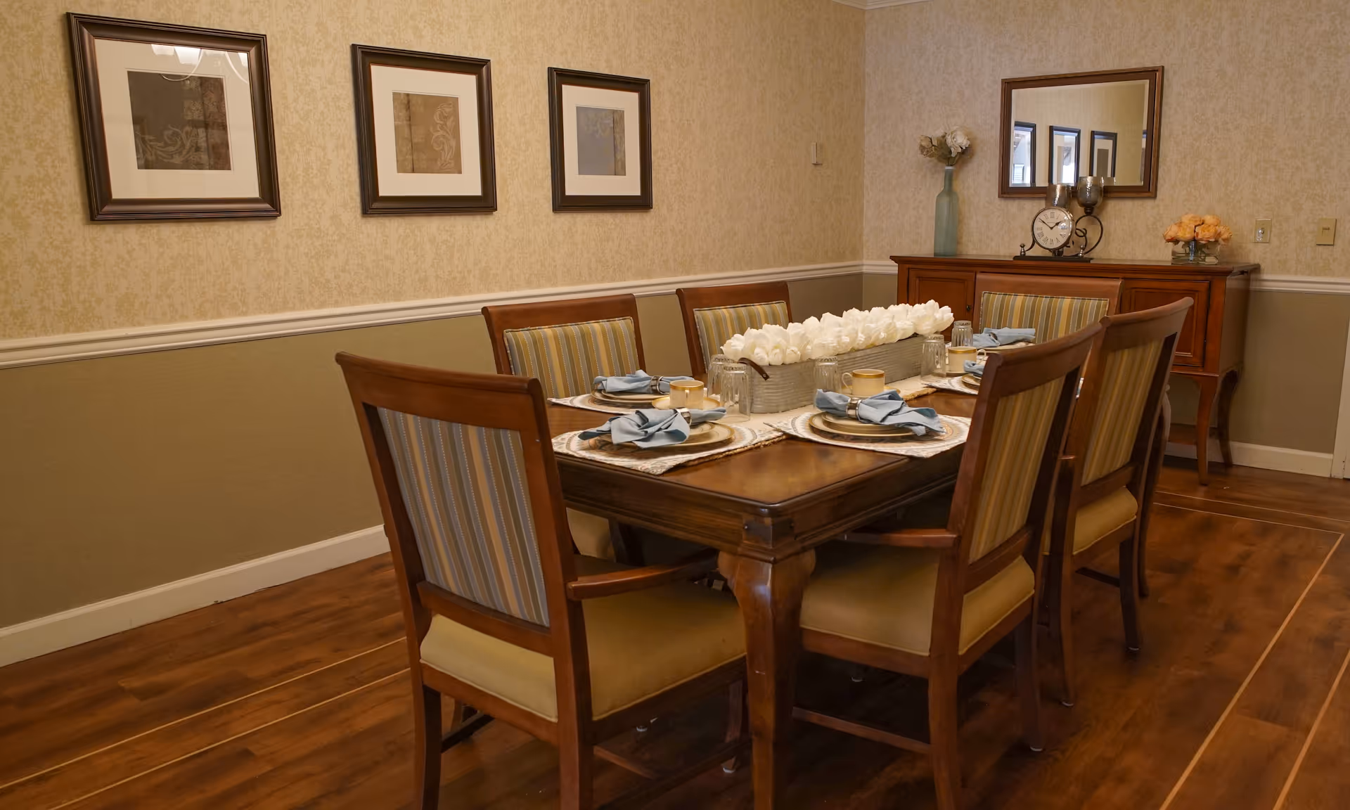 A formal dining room with a wooden dining table set for six people. The table has placemats, plates, cups, and napkins neatly arranged. A long centerpiece with white flowers is placed in the middle of the table. The room has wooden flooring, wallpapered walls with a chair rail, three framed pictures on one wall, and a wooden sideboard with a mirror, clock, and flower arrangements.