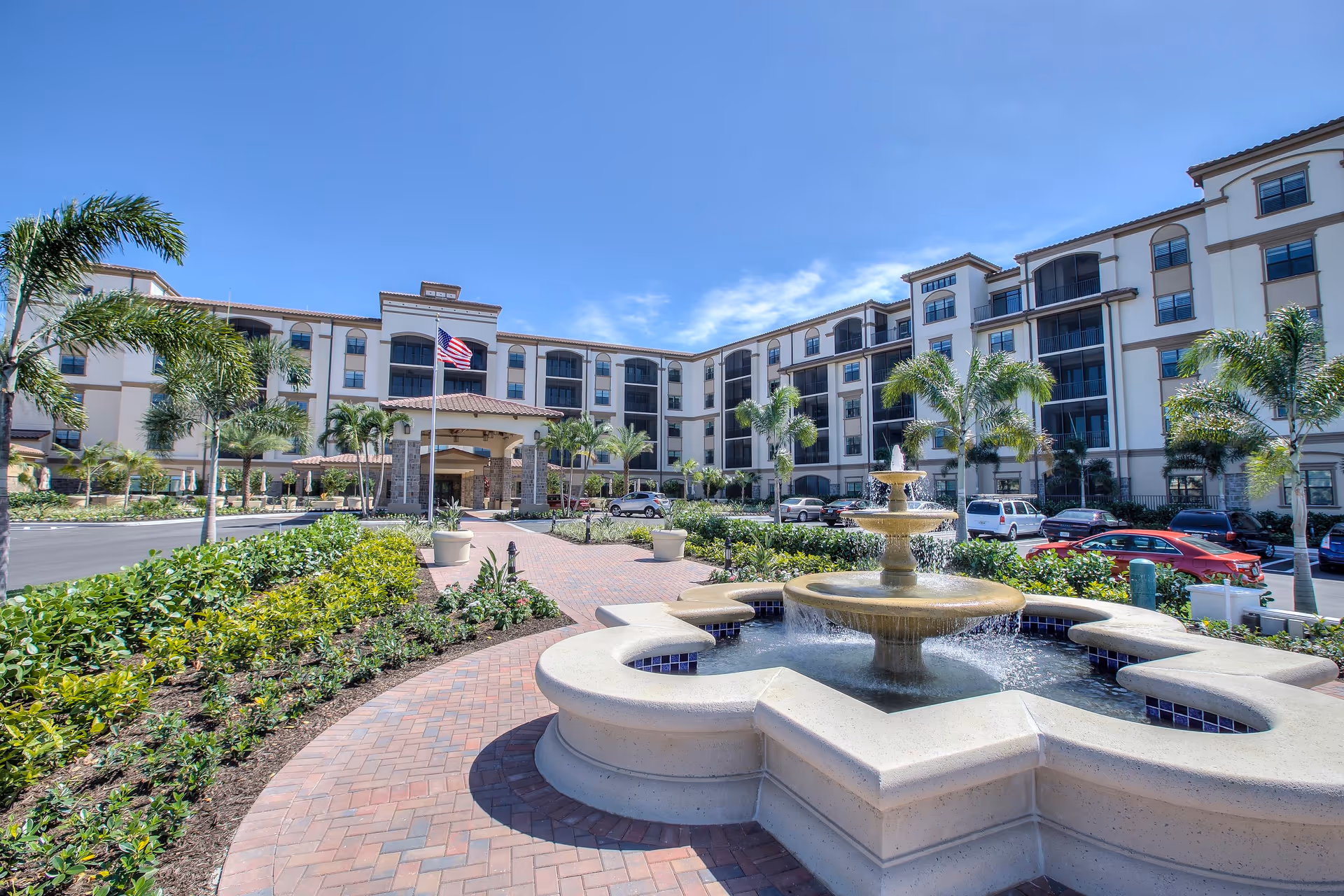 Exterior view of The Arlington of Naples senior living facility showing a large multi-story building with balconies, palm trees, a central fountain, and a paved walkway under a clear blue sky.