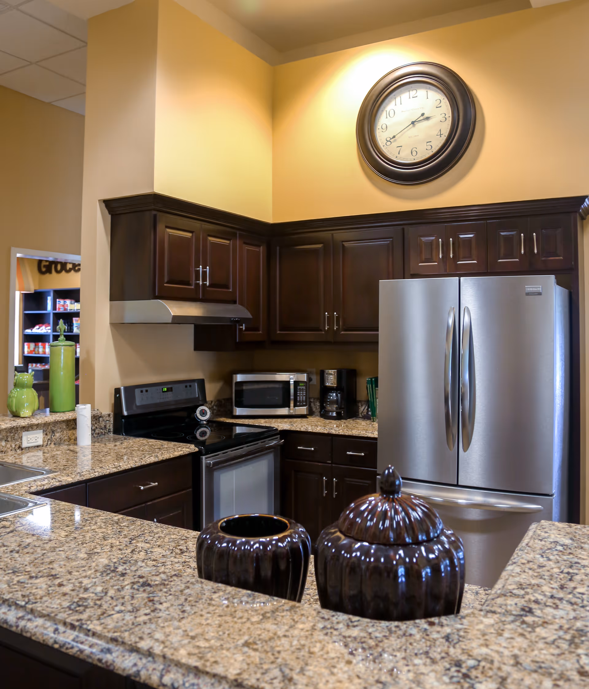 Modern kitchen area with dark wooden cabinets, granite countertops, a stainless steel refrigerator, an electric stove with oven, a microwave, and a coffee maker. A large round clock is mounted on the beige wall above the cabinets. Two decorative dark ceramic jars are placed on the countertop in the foreground.