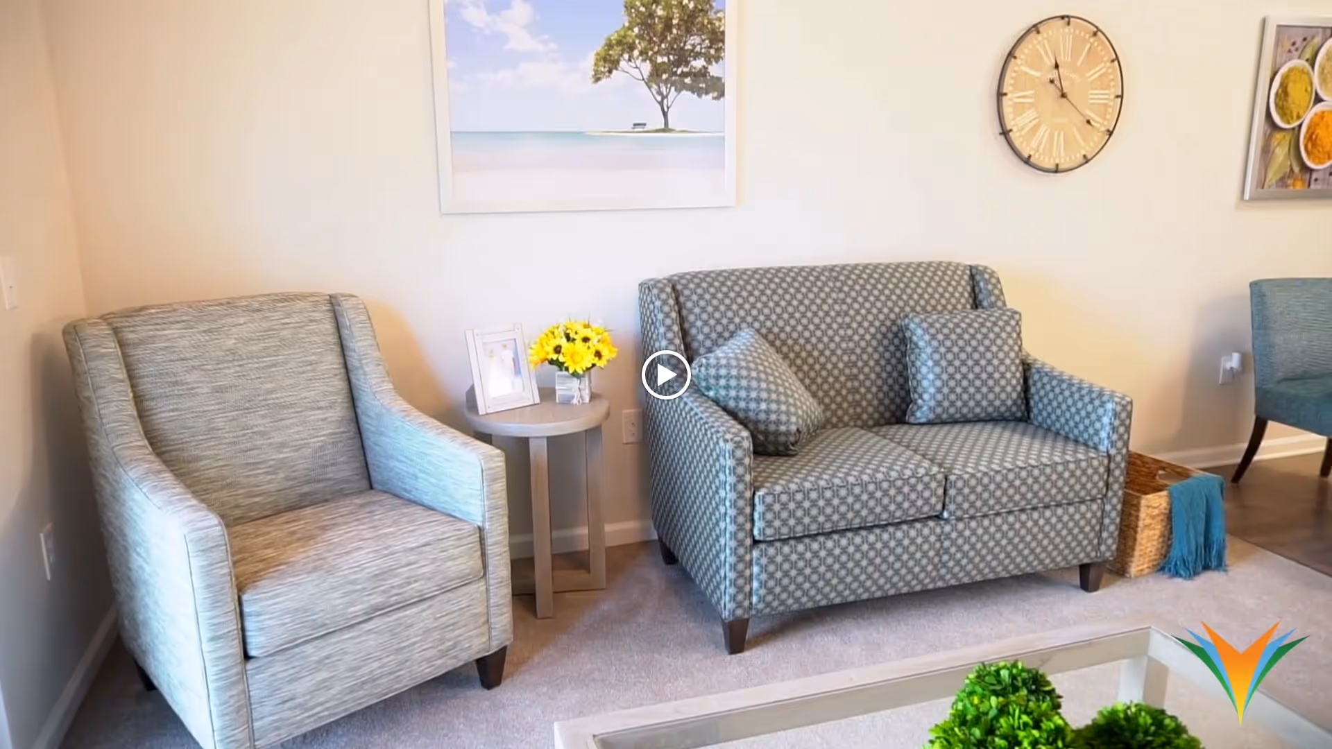 A cozy living room area with a patterned loveseat and matching armchair, a small round side table with a photo frame and a vase of yellow flowers, a wall clock, and framed pictures on the wall. A glass coffee table with green plants is partially visible in the foreground.