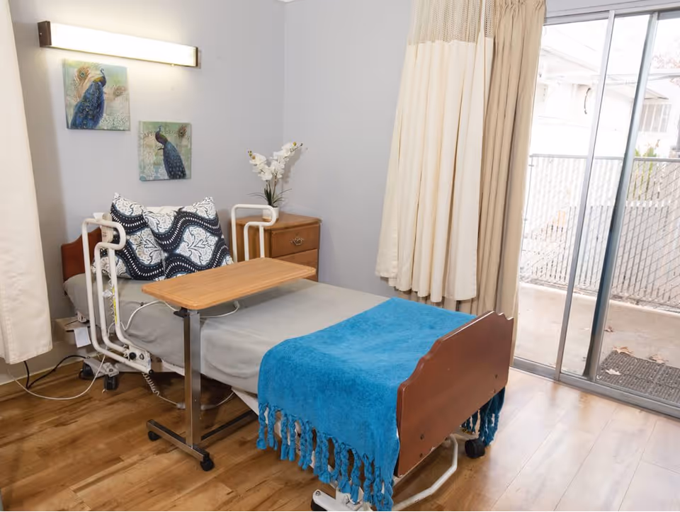 A small, neatly arranged bedroom in a senior living facility featuring a hospital-style bed with white and blue patterned pillows and a blue throw blanket. There is a wooden bedside table with a white orchid plant, two peacock paintings on the wall, a wall-mounted light fixture, and sliding glass doors with beige curtains leading to an outdoor area.