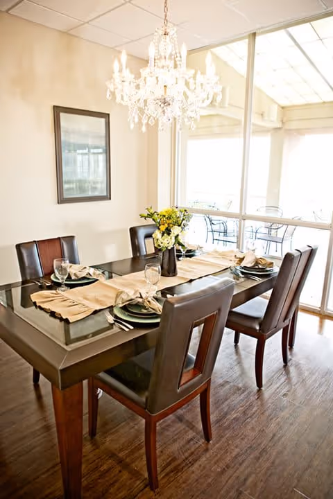 A dining room with a rectangular glass-top table set for six people. The table has beige placemats, plates, napkins, and wine glasses. A vase with yellow and white flowers is in the center. There are six dark brown chairs around the table. A chandelier hangs above the table, and large windows in the background let in natural light, showing an outdoor patio area with metal chairs and tables.