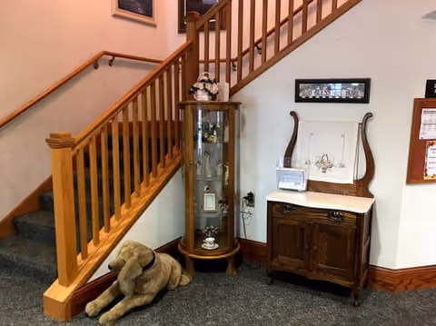 Interior corner of a senior living facility with a carpeted staircase featuring wooden railings. Next to the staircase is a tall glass display cabinet with decorative items inside and a small wooden cabinet with a white top. A large stuffed dog toy is placed on the floor near the stairs. The walls have framed pictures and a bulletin board.
