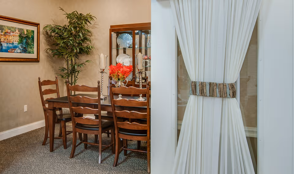 A dining room with a wooden dining table and six matching chairs. The table is decorated with two tall candle holders and a vase with red flowers. Behind the table is a glass cabinet displaying plates and glassware. A large potted plant and a framed painting hang on the wall. To the right, white curtains are tied back with a striped fabric tie.