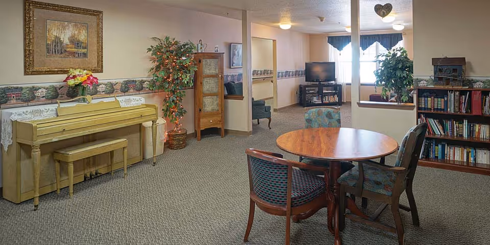 Communal living area with a piano, round table and chairs, bookshelves, TV, and potted plants.