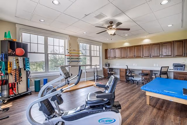 A bright physical therapy or exercise room with large windows, wooden flooring, and a ceiling fan. The room contains exercise equipment including a recumbent bike, parallel bars for walking practice, a blue padded therapy table, and various therapy tools organized on shelves and racks. There are also desks and chairs along the back wall with wooden cabinets above.
