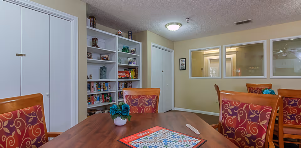 A cozy room with a wooden table and chairs featuring red and gold patterned upholstery. On the table is a Scrabble board game and a small vase with blue flowers. In the background, there is a white bookshelf filled with board games and decorative items, two closed white doors, and two windows looking into another room.