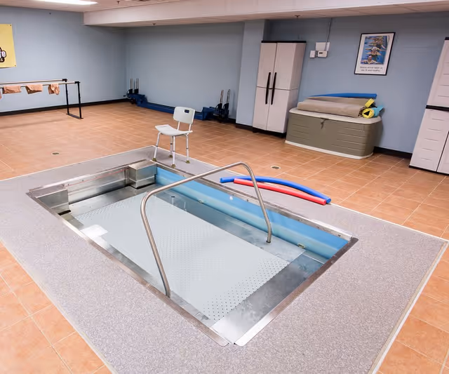 Indoor therapy/hydrotherapy pool with handrails in a tiled room with a chair, storage cabinets, and pool noodles.