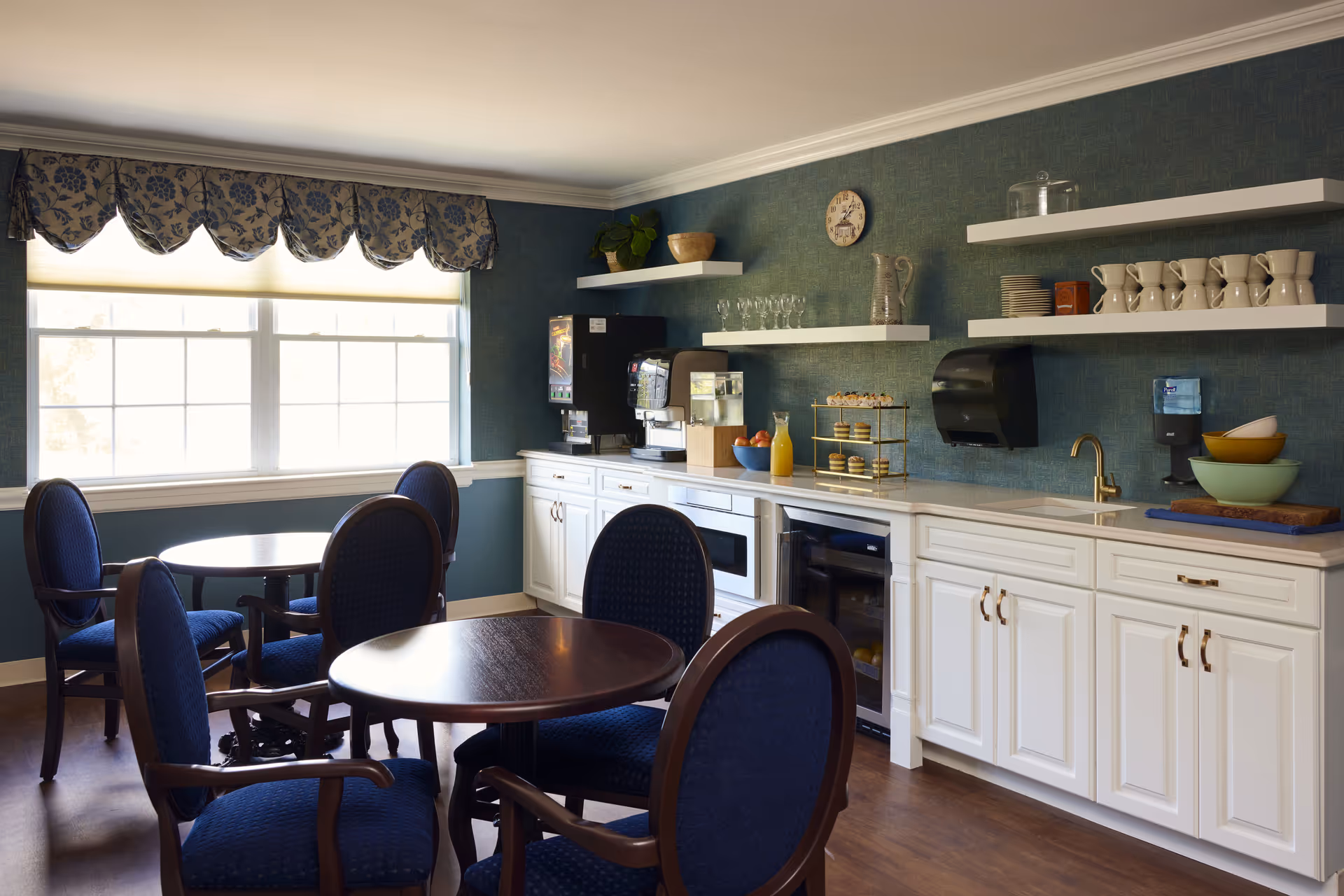 A cozy dining area with round wooden tables and blue upholstered chairs. The room features a large window with floral valance curtains, white cabinetry with a countertop holding a beverage dispenser, glassware, a tiered tray with pastries, and bowls. Shelves above the counter display cups, plates, and decorative items. The walls are covered in textured green wallpaper, and the floor is wooden.