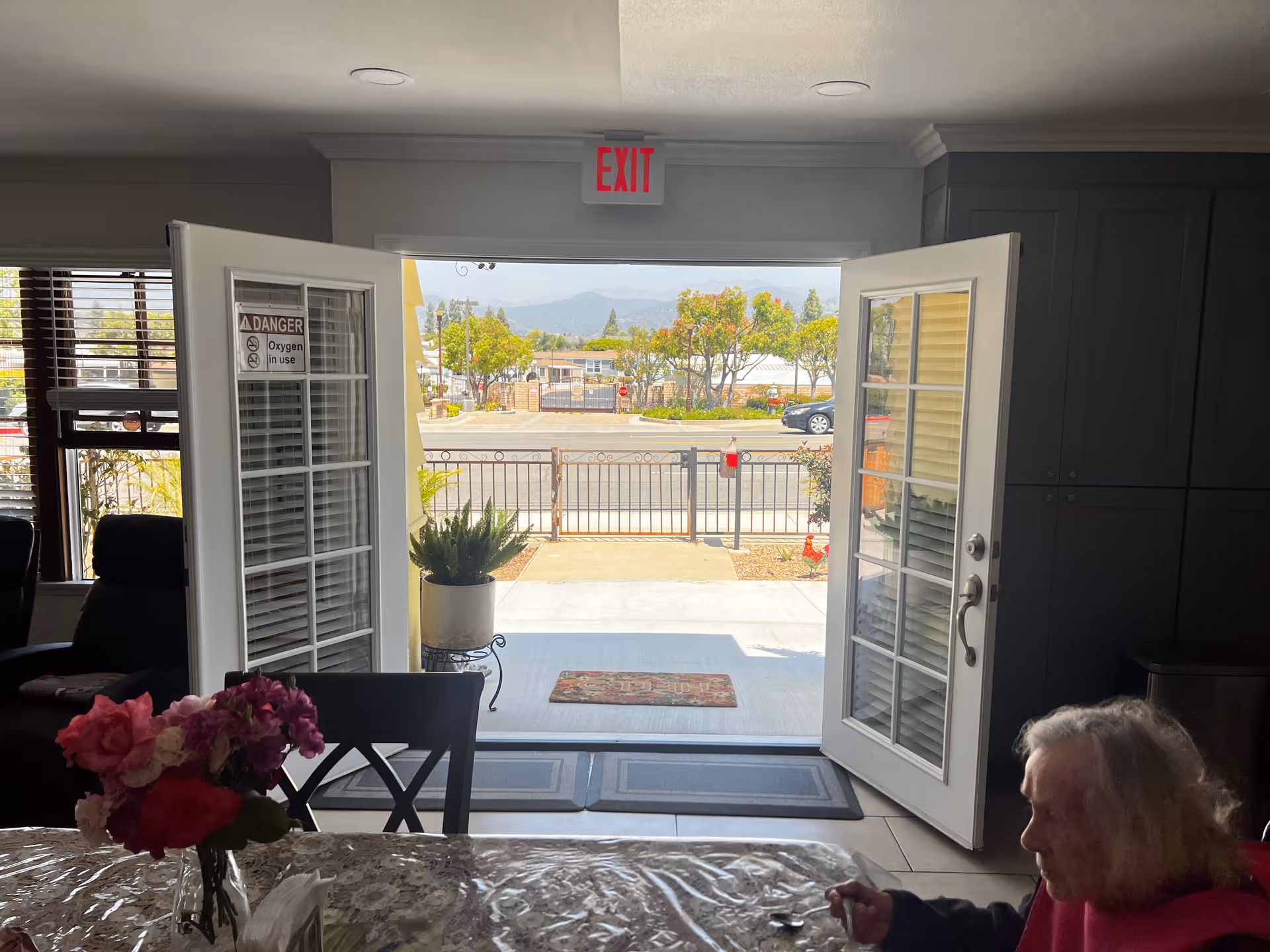 Interior view looking through open double doors to the street, with a table and vase of flowers in the foreground and an elderly woman seated at the right.