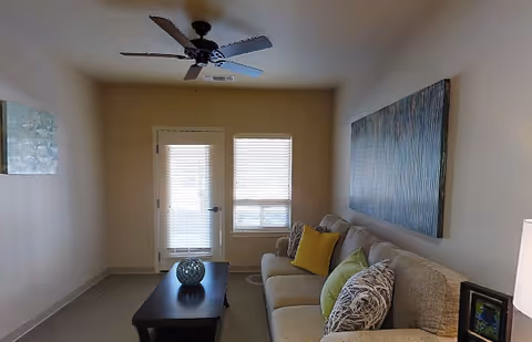 A cozy living room with a beige sofa adorned with multiple colorful pillows, a dark wooden coffee table with a decorative item on it, a ceiling fan with lights, and two windows with blinds allowing natural light to enter. The walls are decorated with two pieces of artwork.