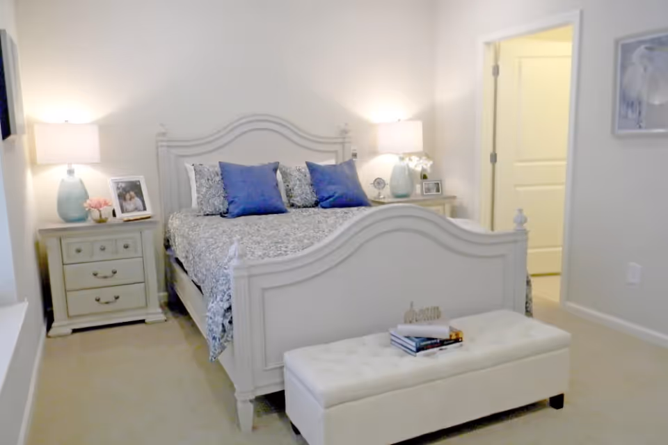 A bright and cozy bedroom with a white wooden bed frame, patterned bedding, and three blue accent pillows. There are two matching nightstands on either side of the bed, each with a lamp and decorative items including framed photos and flowers. A white cushioned bench with books on top is placed at the foot of the bed. The room has beige carpeting and light-colored walls with a doorway leading to another room.