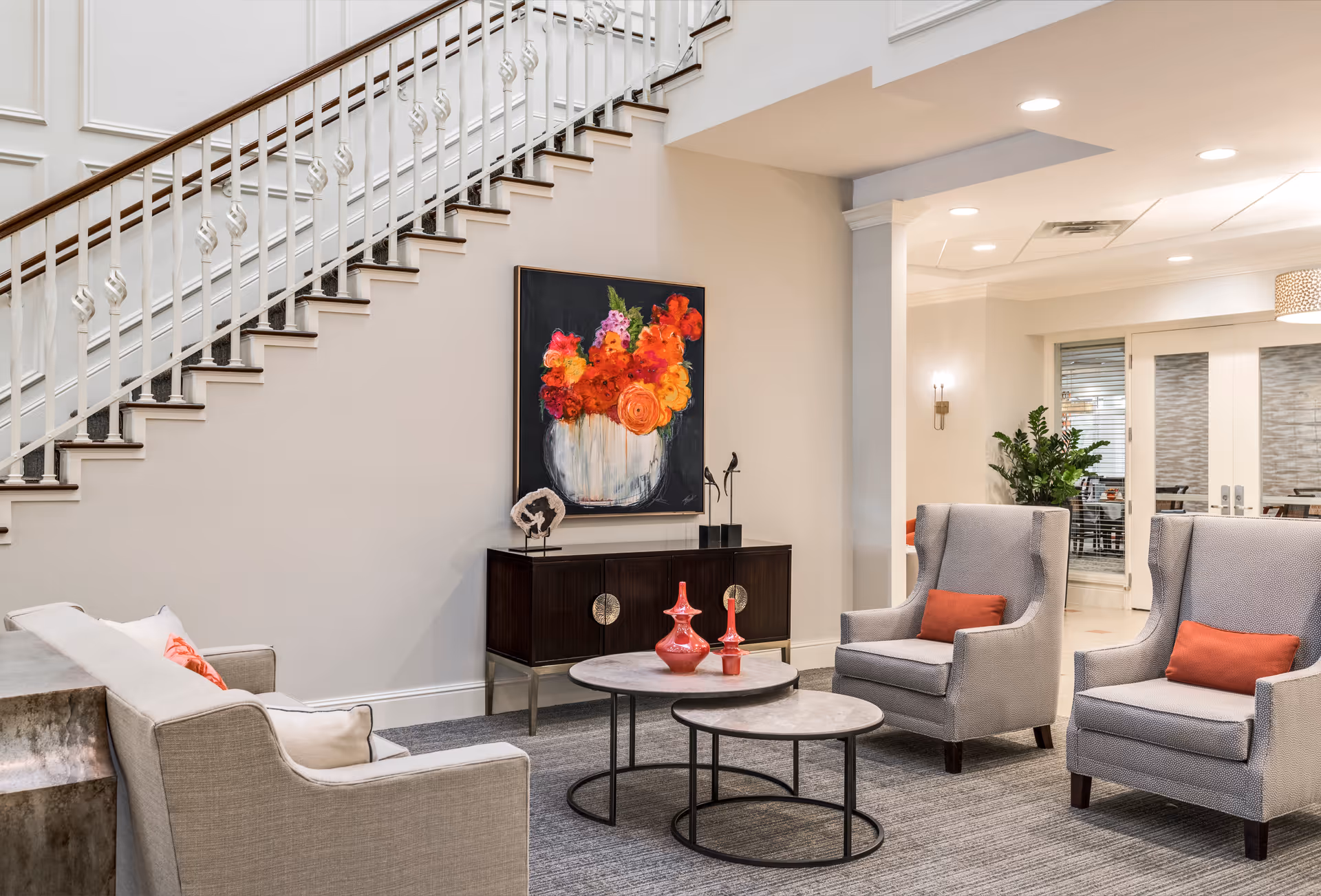 A modern living room area featuring a beige sofa with cushions, two gray armchairs with orange pillows, and two round coffee tables with decorative red vases. Behind the seating area is a dark wooden cabinet with decorative items and a colorful painting of flowers in a vase hanging on the wall. A staircase with white railings is visible on the left side, and a doorway leading to another room is on the right.