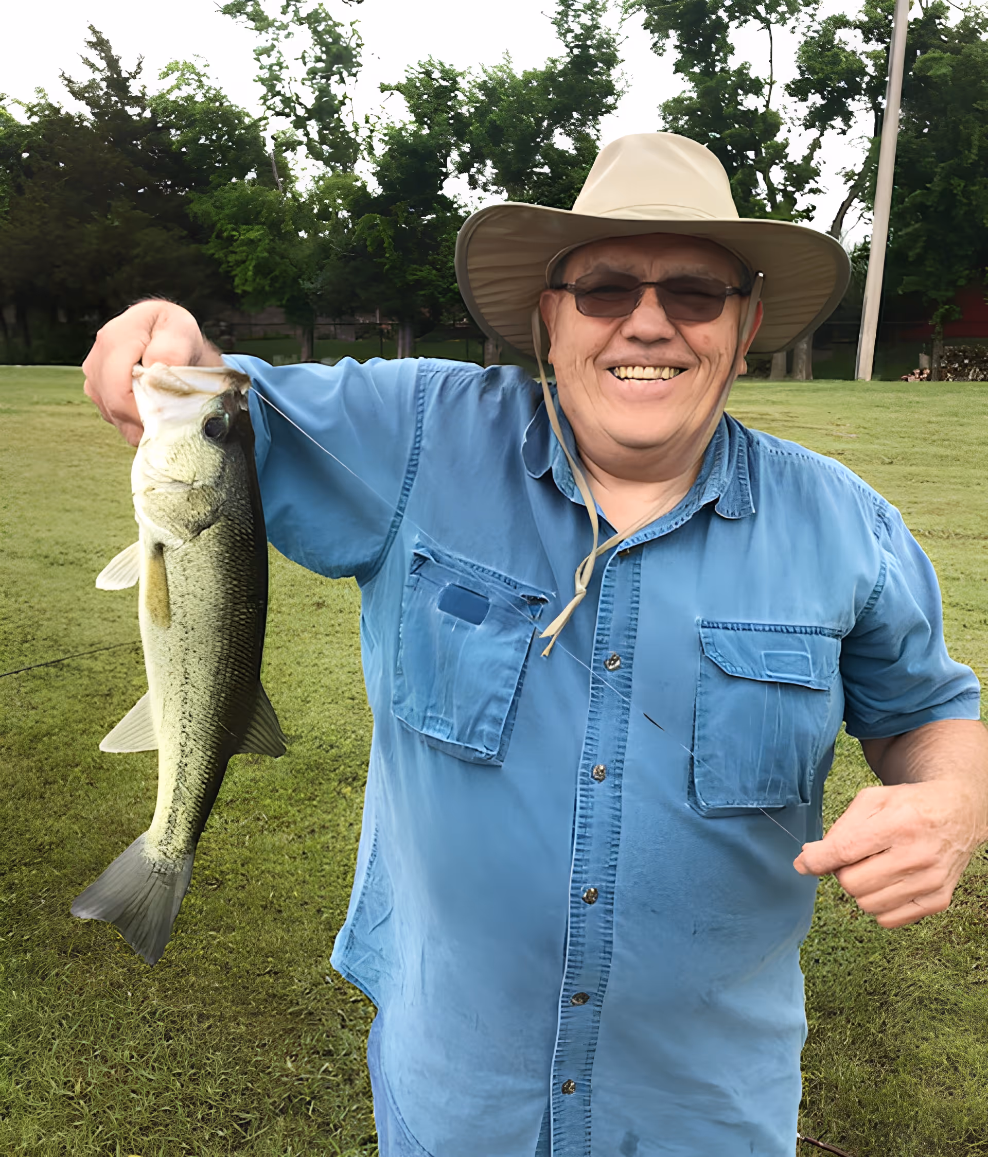 An elderly man wearing a wide-brimmed hat and sunglasses smiles while holding a large fish in one hand, standing on a grassy area with trees in the background.