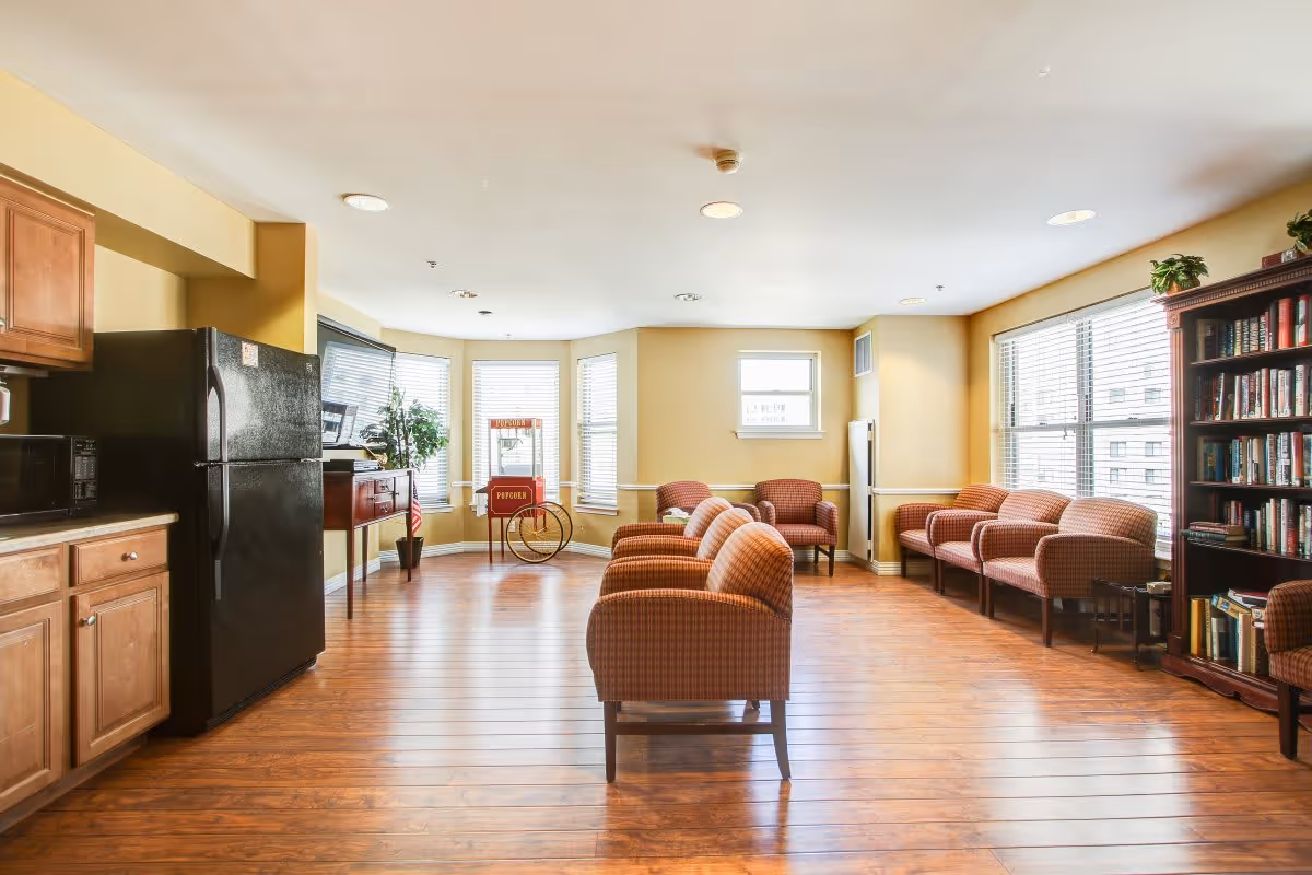 A bright and spacious common area with wooden flooring, featuring a row of cushioned chairs in the center and along the walls. There is a black refrigerator and wooden cabinets on the left side, a popcorn machine near the windows, and a tall bookshelf filled with books on the right. Large windows allow natural light to fill the room.