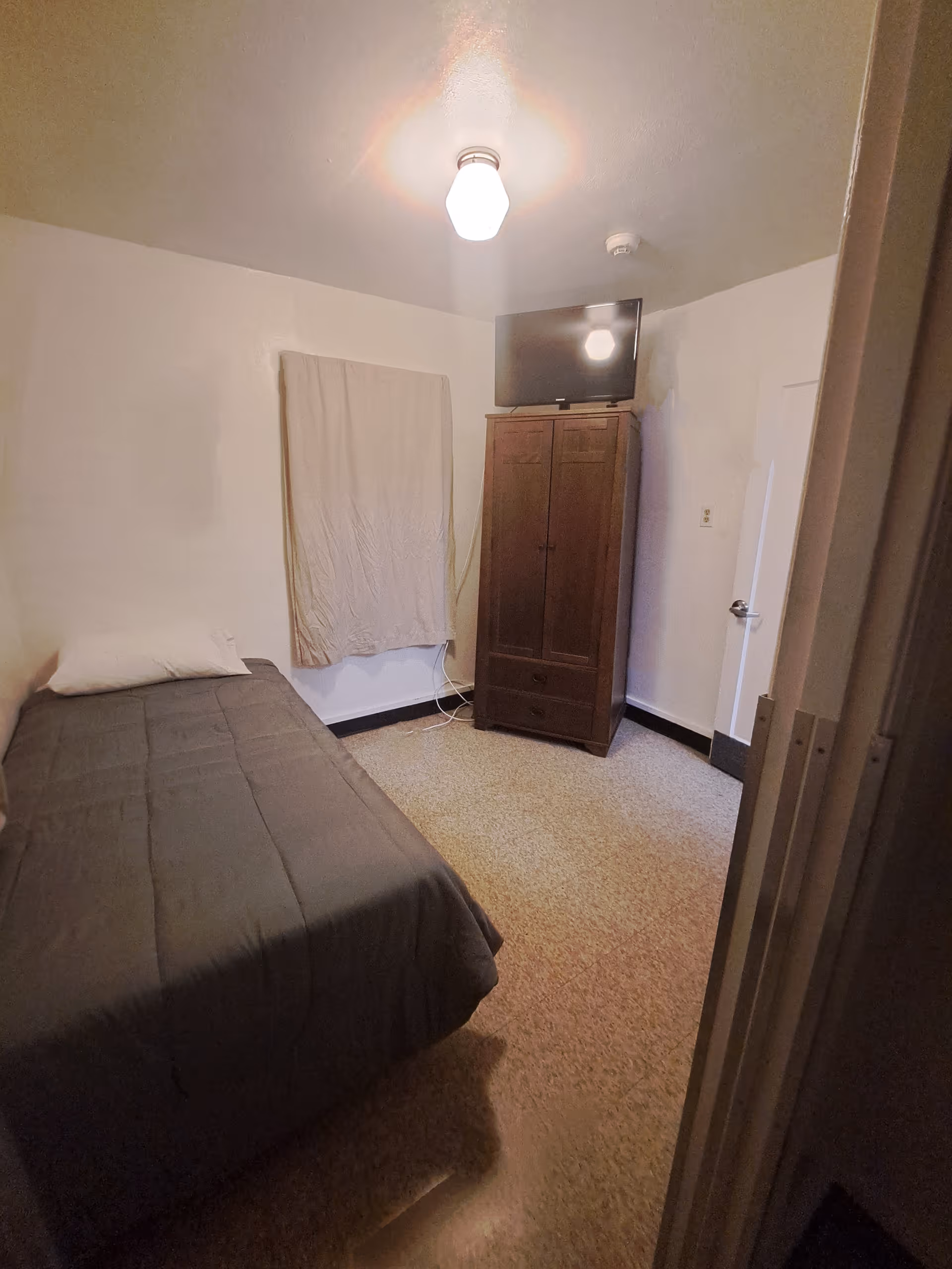 A small bedroom with a single bed covered in a dark gray blanket and a white pillow. There is a wooden wardrobe with a flat-screen TV on top against the far wall. A window is covered with a beige curtain. The room has beige carpet flooring and white walls, with a ceiling light fixture turned on.