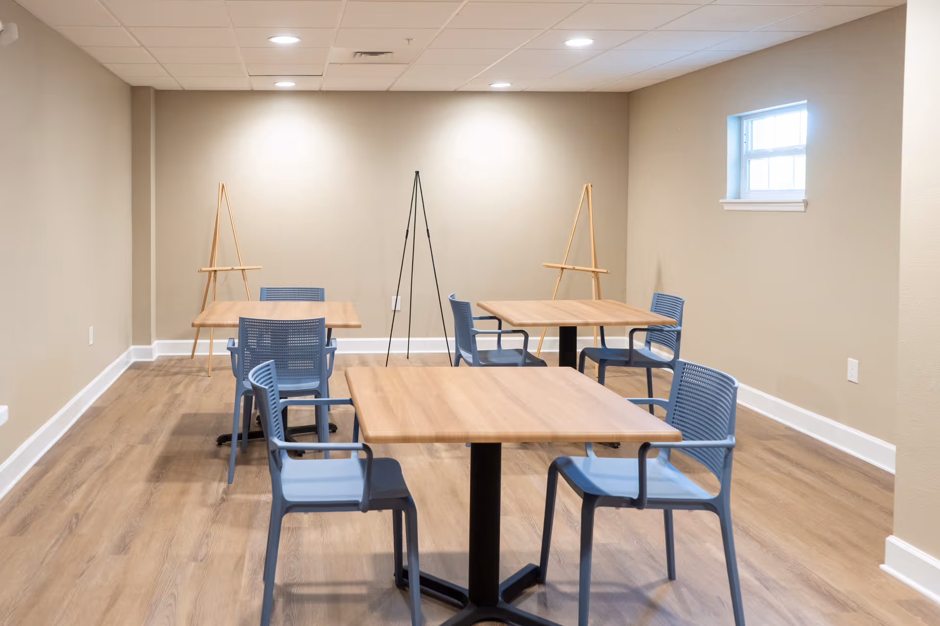 A small room with three wooden tables, each surrounded by blue plastic chairs. The room has light beige walls, wood flooring, a small window on the right wall, and two wooden easels and one black easel stand against the back wall.