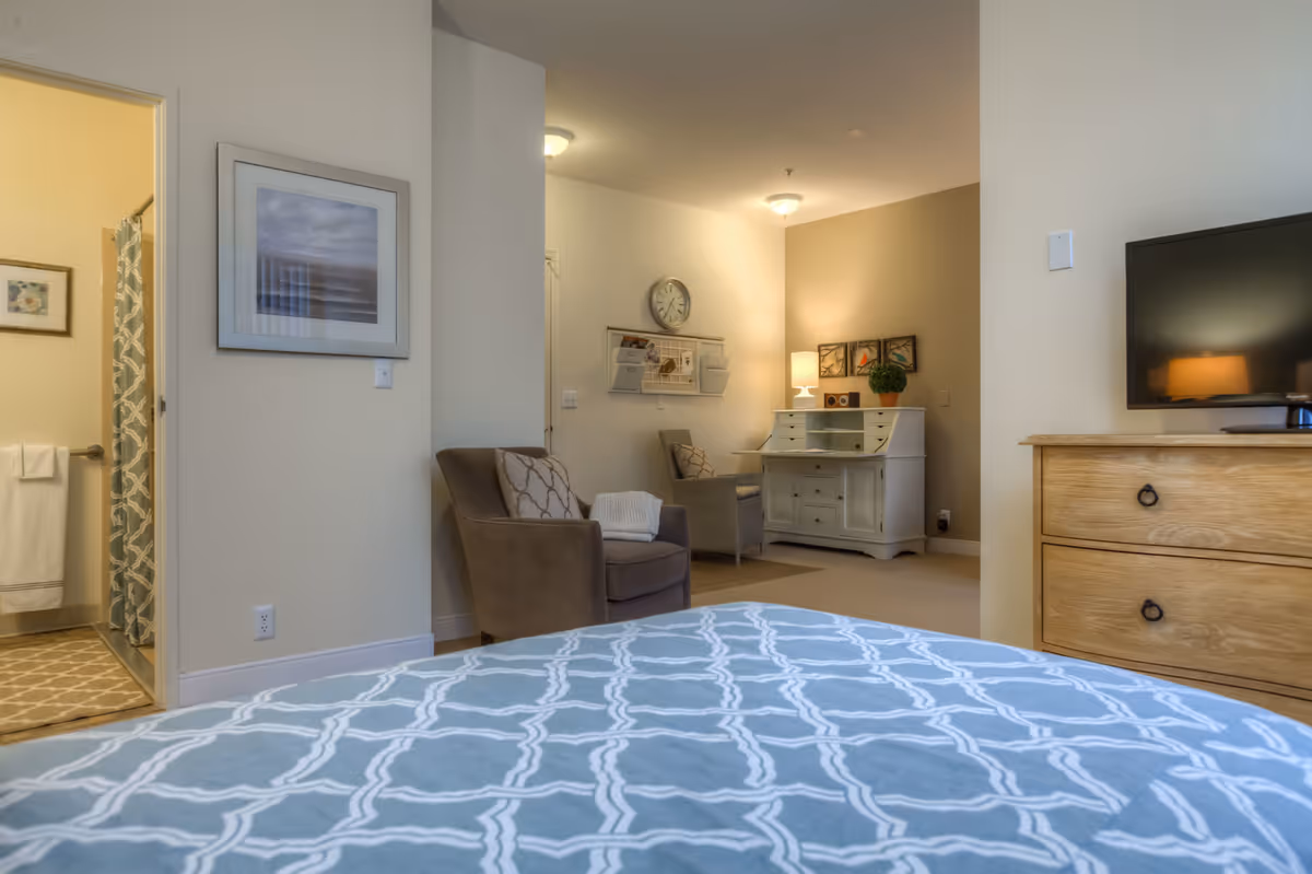 Bright bedroom with a patterned bedspread in the foreground, a TV on a wooden dresser, a chair and desk seating area, and an open bathroom door.