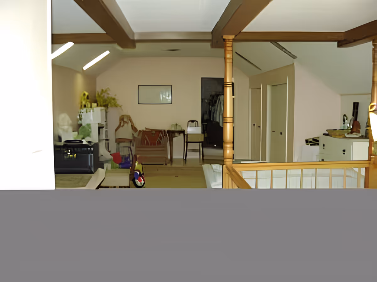 Interior view of a room with wooden beams on the ceiling, various chairs, a table, and shelves with plants and other items. There is a railing in the foreground and doors along the right wall.