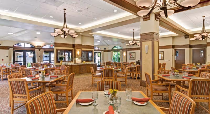 A spacious dining room in Merrill Gardens at Campbell featuring multiple tables with striped cushioned chairs. Each table is set with glasses, plates, and red napkins. The room has large windows with arched tops allowing natural light to enter, beige walls with wooden trim, and elegant ceiling lights.