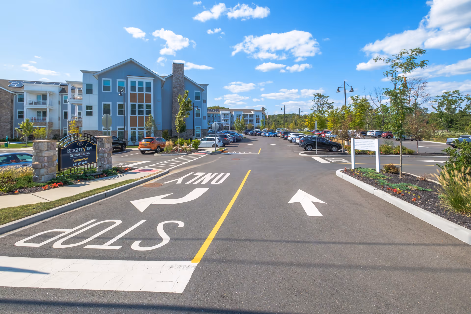 View of the parking lot and exterior of Brightview Eatontown senior living facility on a sunny day with blue sky and scattered clouds. The building is modern with multiple floors, large windows, and stone accents. There are cars parked along the sides of the lot and landscaped areas with trees and flowers.