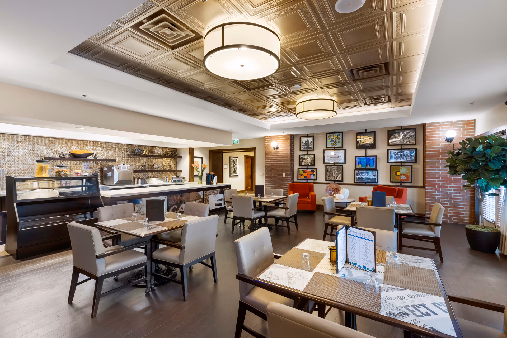 Bright, modern dining room with multiple tables and chairs, a service counter, decorative ceiling lights, and framed wall art.