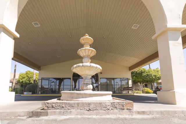 A three-tiered stone fountain is centered under a large covered entrance with an arched ceiling. The building exterior is light-colored with large windows and some greenery visible in the background.