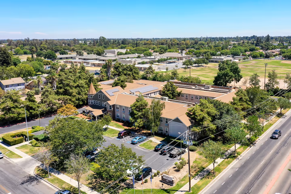 Aerial view of Pacifica Senior Living Merced facility surrounded by trees and greenery, with a parking lot and several cars visible. The building has a tan roof and light-colored exterior walls, situated near a main road and open grassy areas.