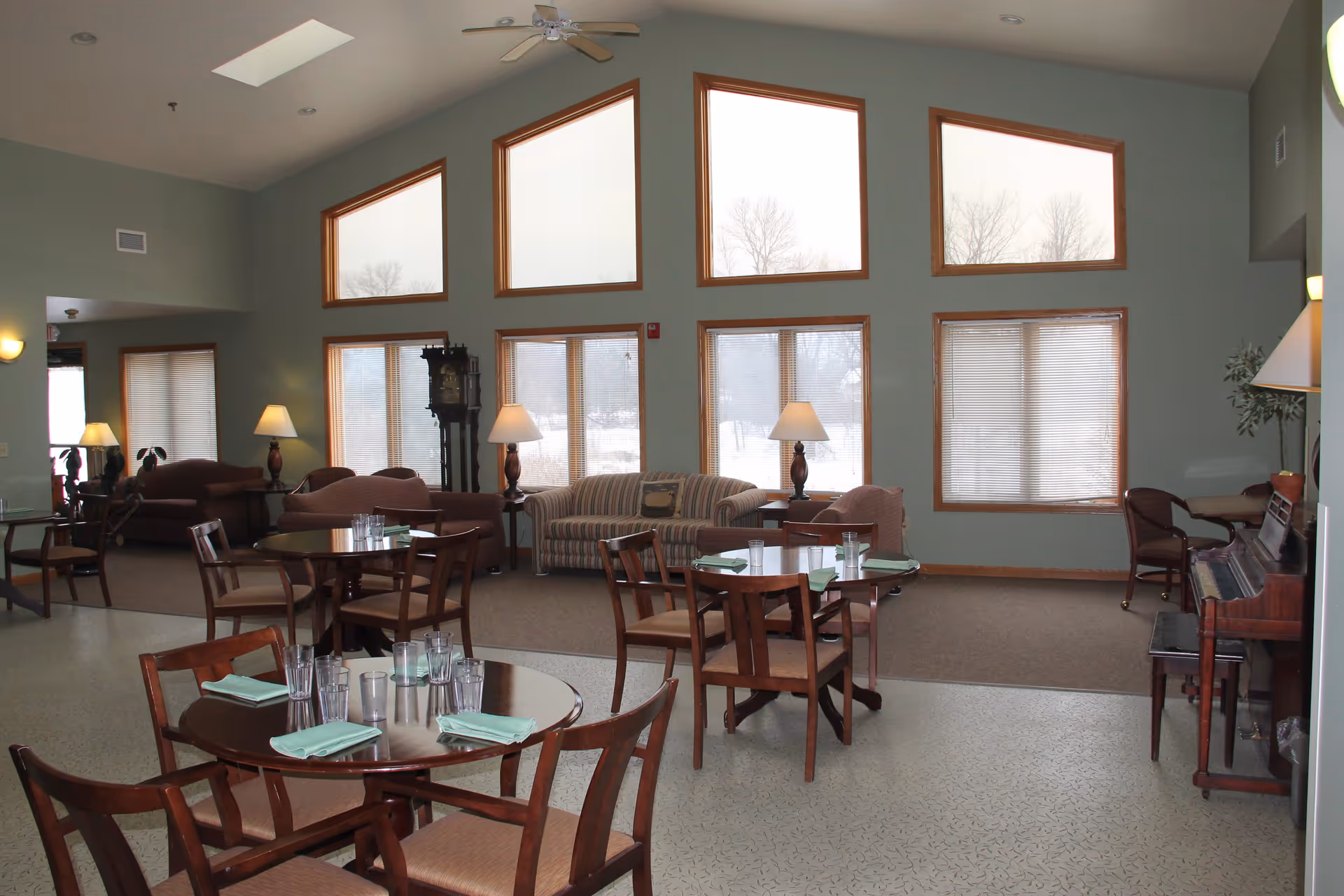 A spacious common area with multiple round wooden tables and chairs set with glasses and folded napkins. The room has large windows with wooden frames, some with blinds, and high triangular windows near the ceiling. There are several sofas and armchairs arranged along the walls, with table lamps and a grandfather clock. A piano is visible on the right side of the room. The walls are painted light green and the floor has a patterned linoleum surface.